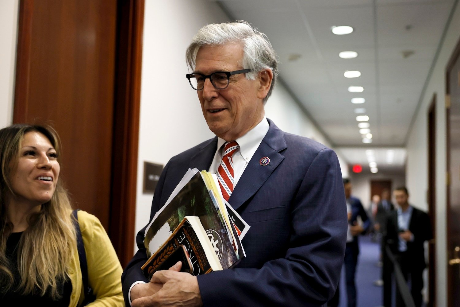 Congressman Don Beyer of Virginia walking through an office, wearing a suit and holding papers. To the left is a women looking at him smiling.