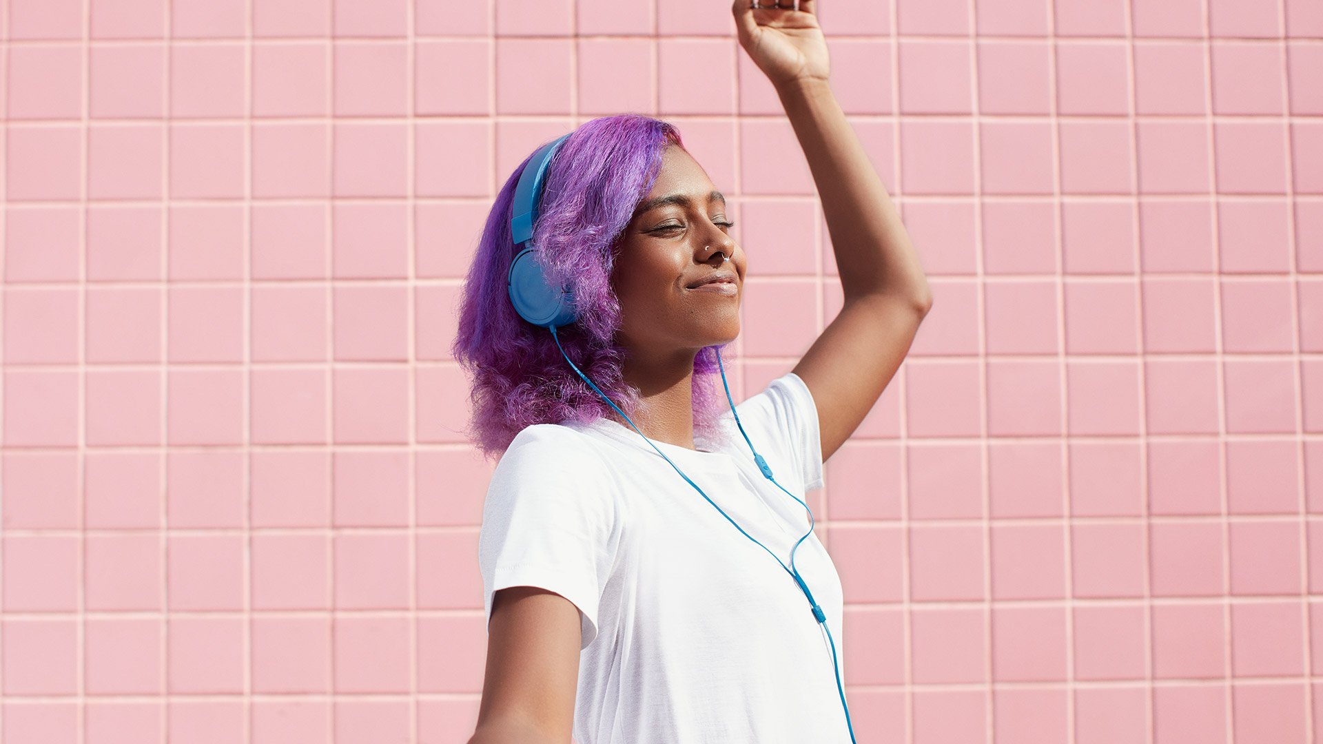 Woman with right hand raised with lilac hair wearing a pair of light blue headphones against a pink tiled wall